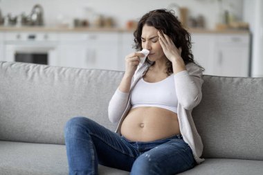 Portrait Of Sick Young Pregnant Lady Feeling Unwell At Home, Ill Expectant Female Blowing Runny Nose In Tissue And Touching Head, Suffering Seasonal Flu And Migraine, Sitting On Couch In Living Room