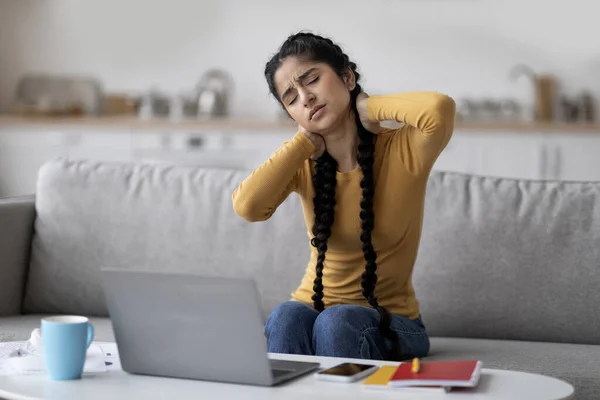 Tired Indian Woman Suffering Neck Pain While Working Or Studying With Laptop Computer At Home, Exhausted Young Hindu Female Sitting On Couch And Massaging Painful Area, Suffering Osteoporosis