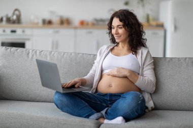 Smiling young pregnant woman relaxing with laptop on couch at home, beautiful expectant lady browsing internet, shopping online, or reading pregnancy blog while sitting on sofa in living room