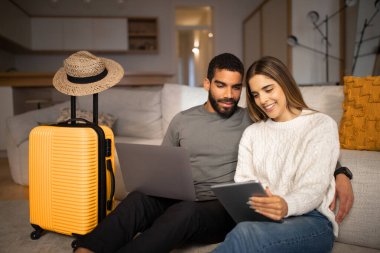 Smiling young arab male, european lady use tablet and laptop to choose country for vacation travel, buy tickets in living room interior with suitcase, hat. Travel app for tourists, love relationship