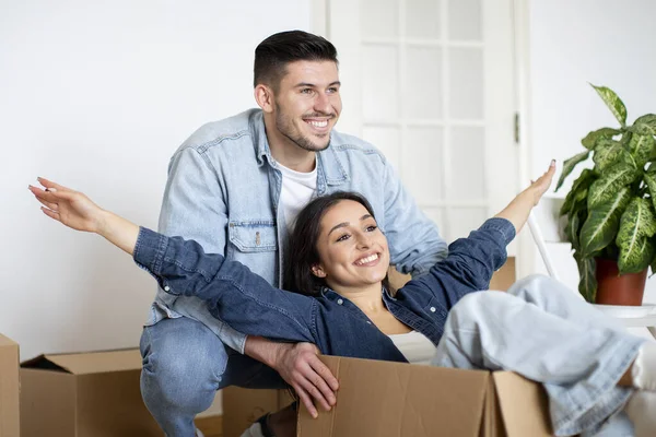 Excited young spouses having fun on moving day, closeup shot of cheerful man pushing cardboard box with his happy wife riding in container, celebrating relocation into new home, free space