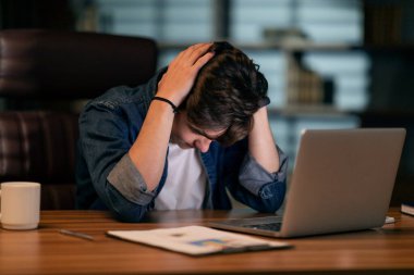 Burnout at work, overworking concept. Exhausted young man sitting at workplace in front of computer at dark office, touching his head, lack of ideas, looking for creative business solution, copy space