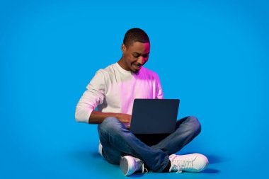 Positive handsome young black man in casual sitting on floor, using modern laptop over blue background in neon light, websurfing or working online, copy space. Modern technologies concept