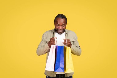 Glad shocked middle aged black male shopaholic in casual looking in package with purchases, enjoy fashion isolated on yellow background, studio. Huge sale, shopping, gift buying, new clothes emotions