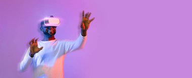 Augmented reality concept, web-banner. Black man in white using wireless VR headset on neon studio background, touching something invisible with hands, panorama with copy space, blurred