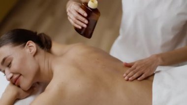 Professional spa service. Close up shot of unrecognizable masseur pouring warming oil on female client back and massaging her, tracking shot, slow motion, free space