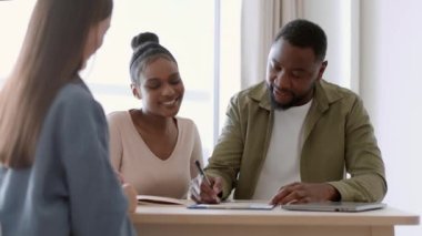 Great family purchase. Young happy african american couple signing mortgage agreement and getting keys from agent, embracing at real estate agency, tracking shot, slow motion, free space