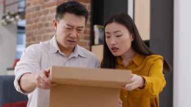 Wrong package. Couple of asian spouses opening long awaited delivery box, looking inside and feeling displeased, disappointed with bad purchase at home