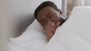 Night symptoms of sickness. Close up portrait of sleeping african american guy waking up and coughing, keep resting in his bed, tracking shot, slow motion, free space