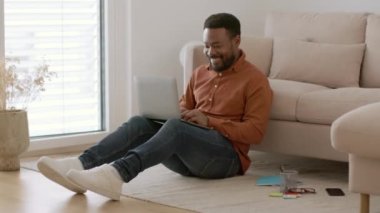 Online leisure concept. Young positive african american guy playing video games on laptop, resting on floor at home on weekends, tracking shot, slow motion, empty space
