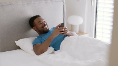 Happy morning. Young positive black guy web surfing on smartphone, waking up at home, resting in bed on weekend, tracking shot, slow motion, empty space