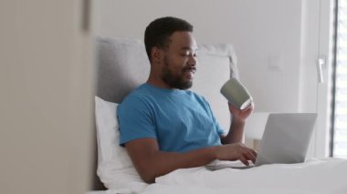 Cozy morning on freelance. Young happy african american guy manager working on laptop in bed and drinking his coffee, enjoying his lifestyle, tracking shot, slow motion, free space