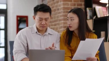 Domestic accounting issues. Asian spouses filing tax form on laptop and comparing expenses on paper bills, having some financial problems at home, tracking shot, slow motion