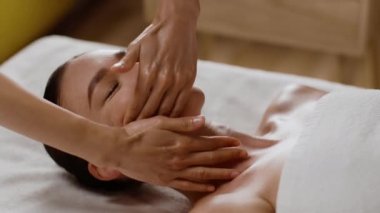 Stress relief and rejuvenation concept. Relaxed woman lying on spa bed for facial and head massage, enjoying treatment by masseur at luxury wellness resort, close up portrait, slow motion