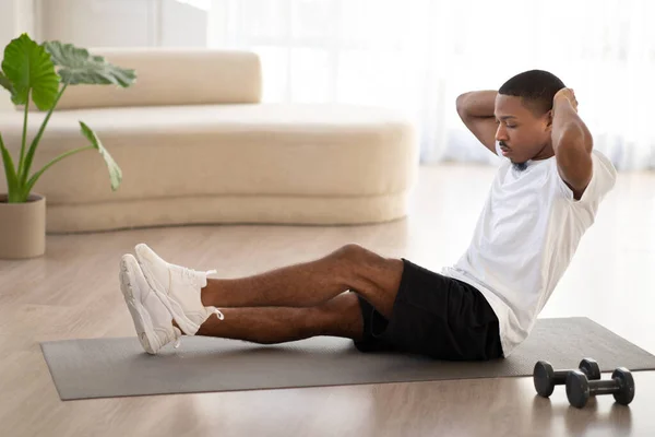 Motivated athletic handsome young black man in sportswear sitting on yoga mat, doing crunches for abs workout at home, using fitness tools, looking at copy space, full length candid shot