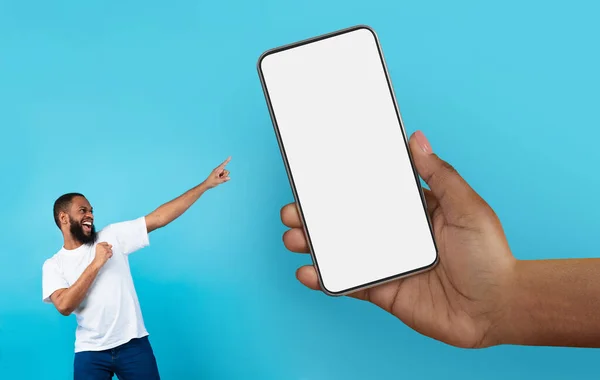 Online Offer. Cheerful Black Man Pointing At Huge Hand With Blank Smartphone, Excited African American Man Demonstrating Copy Space For Mobile Advertisement While Standing On Blue Background, Mockup