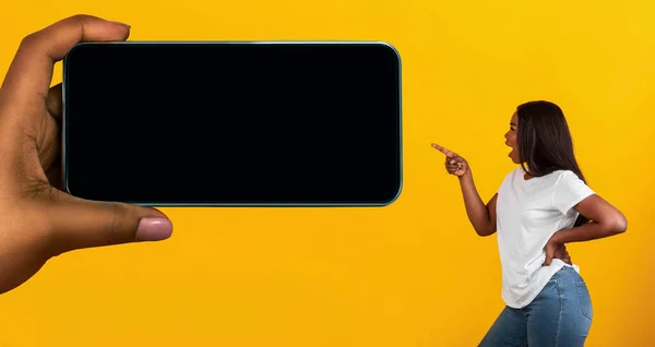 Excited Black Woman Pointing Aside At Huge Female Hand With Blank Smartphone On Yellow Background, Surprised African American Lady Demonstrating Copy Space For Mobile App Or Website Design, Mockup