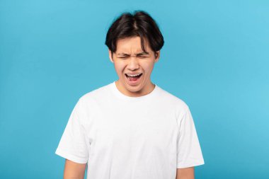 Sad Asian Teen Guy Shouting Loudly And Crying Expressing Negative Emotions Standing Over Blue Studio Background. Portrait Of Teenager Boy Screaming Having Nervous Breakdown Problem