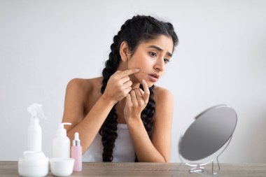 Skin Problems. Upset Young Indian Woman Looking At Pimple On Her Cheek, Sad Beautiful Hindu Female Sitting Near Magnifying Mirror At Home And Inspecting Her Face, Suffering Acne, Copy Space