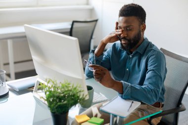 Exhausted black businessman taking off eyeglasses, feeling tired due to computer overwork, sitting at workpalce in office. Stressed african american man suffering from eyes strain