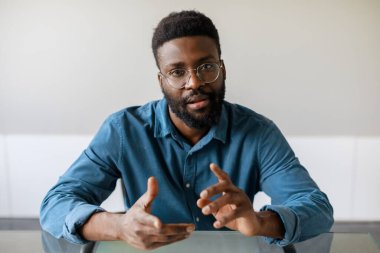 Serious black businessman having video call, talking to camera and gesturing, discussing business project, sitting in office. Entrepreneurship and communication concept