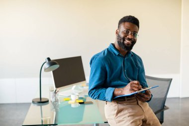 Successful entrepreneurship concept. Excited black businessman checking financial papers while standing near desk in office, happy manager holding clipboard with documents and smiling, copy space