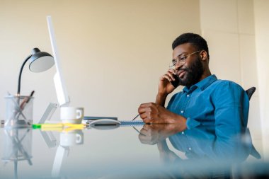 Black businessman working in office and talking on cellphone consulting client or customer online, sitting at workplace. Happy male entrepreneur having smartphone consultation with partner