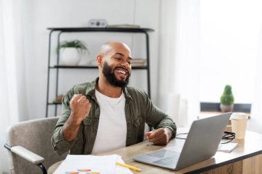 Great job. Happy latin man looking at laptop and expressing emotions of victory, sitting at desk at home office and rejoicing success, free space
