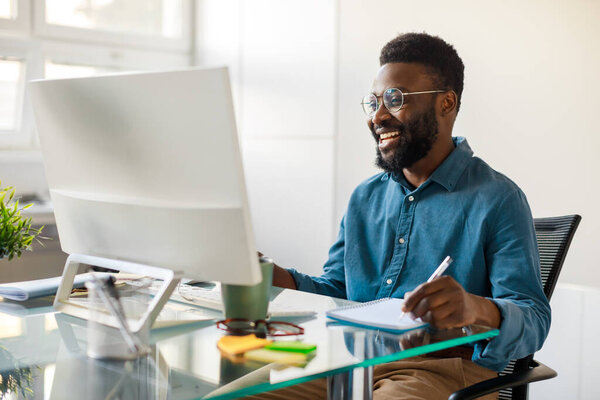 Positive black businessman writing notes in notebook while watching webinar video course, looking at computer monitor and smiling, sitting at workplace in office