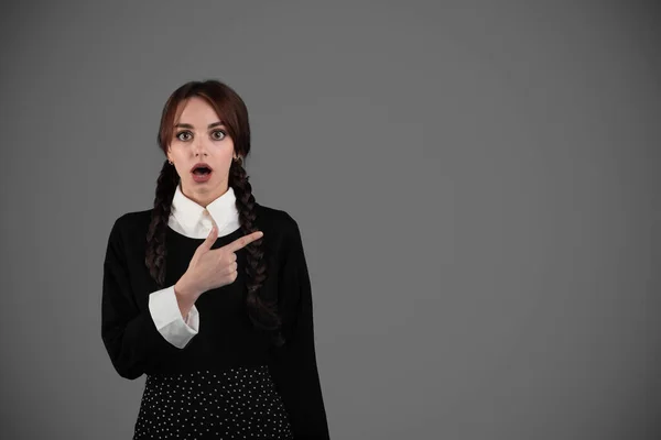 Shocked young female student in gothic black clothes with pigtails with open mouth point finger at free space, isolated on gray wall studio background. Emotions at news, surprise at study, education