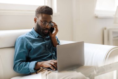 Focused black male entrepreneur talking on smartphone while sitting on sofa and using laptop, free space. Confident businessman looking at laptop screen, working in corporate office