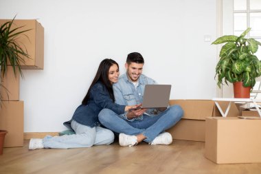 Moving Concept. Smiling Young Couple Using Laptop While Sitting On Floor In Apartment Among Cardboard Boxes, Millennial Spouses Shopping Online After Relocation To New Home, Copy Space