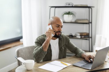 Business communication and entrepreneurship concept. Latin male freelancer calling by cellphone while working online on laptop and communicating, sitting at desk at home office.