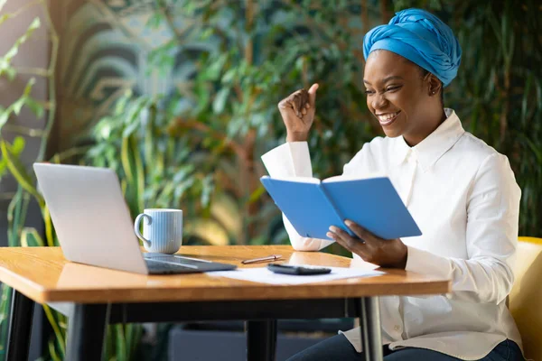 Emotional happy young black lady independent contractor working online at cafe, sitting at table, looking at laptop screen, holding notepad, clenching fist and smiling, enjoying success, copy space