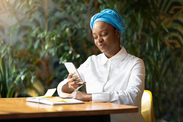 Happy attractive young black woman in smart casual working nomad SMM manager sitting at cafe alone, checking her notepad and using smartphone, chatting with clients, copy space. Remote job concept