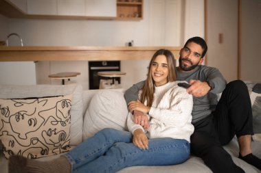Smiling young arab man hugging european female, watching tv with remote control, enjoy romantic date and free time, sit in living room interior. Love, relationship, fun together and movie evening