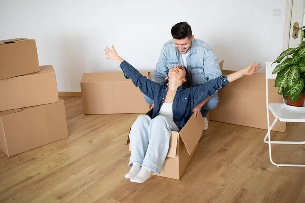 Young Man Riding His Happy Wife In Cardboard Box On Moving Day, Cheerful Young Couple Fooling In Their New Home, Happy Spouses Having Fun While Unpacking Things During Relocation, Copy Space