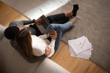 Young arab man hugging european wife with coffee cup, typing on computer, sit on floor in living room interior with documents and graphs. Love, relationship, business, work remotely together at home