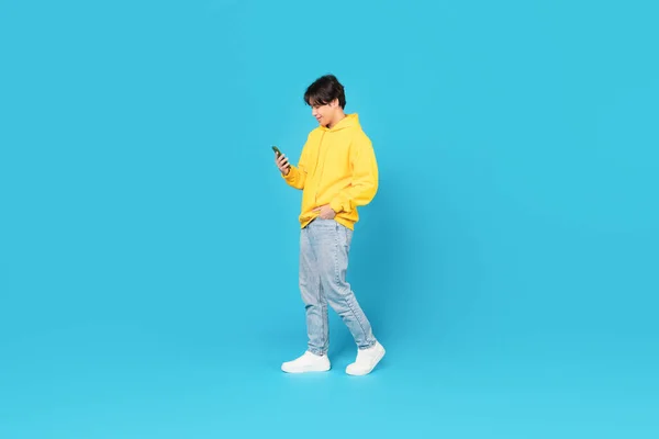 Asian Teen Boy Using Smartphone Walking Posing On Blue Studio Background. Shot Of A Guy Playing Games And Communicating Online Via Phone. New Mobile Application Advertisement. Full Length