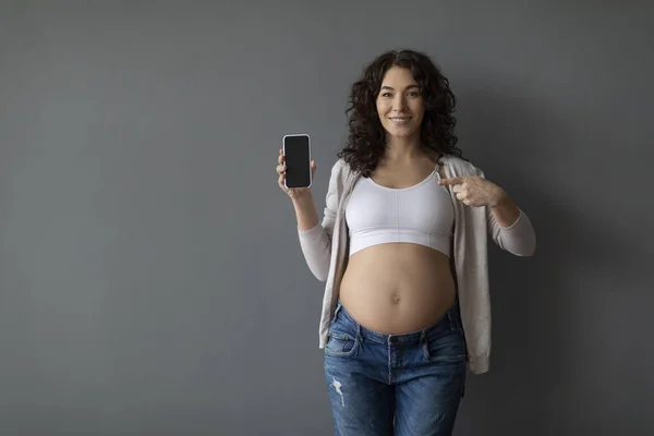 Great App. Smiling Pregnant Lady Showing Blank Smartphone And Pointing At It, Happy Young Expectant Woman Advertising Modern Mobile Application For Pregnancy Tracking, Grey Background, Mockup