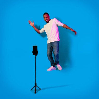 Famous african american influencer cool millennial guy recording content for social media, looking at cell phone set on tripod, dancing and smiling on blue background in neon light, full length