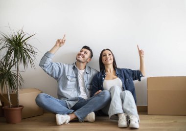Excited Young Spouses Sitting On Floor Among Cardboard Boxes And Pointing Up, Cheerful Couple Having Design Idea After Moving Home, Dreaming About Interior In New Apartment, Copy Space