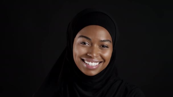 イスラム教徒の美しさ 若いですかなりアフリカ系アメリカ人のイスラム教徒の女性の肖像画を閉じます伝統的なヒジャーブを身に着けていますカメラに笑顔 黒スタジオの背景 スローモーション — ストック動画