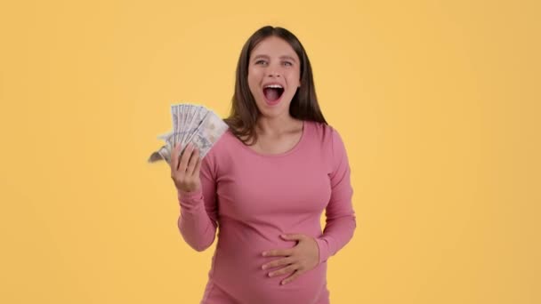 产妇津贴 年轻兴奋的孕妇拿着钱大喊大叫 橙色背景 慢动作 自由自在空间的工作室肖像 — 图库视频影像