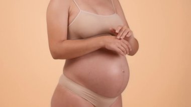 Spa during pregnancy. Close up shot of unrecognizable pregnant lady in underwear rubbing cream on her hands, beige studio background, slow motion, free space