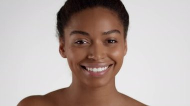 Skin nourishing concept. Close up portrait of young positive african american woman applying serum on face and rubbing it, white studio background, slow motion
