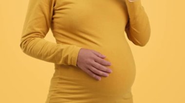 Pregnancy and alcohol. Close up shot of unrecognizable pregnant woman drinking red wine and caressing her belly, orange studio background, slow motion, free space