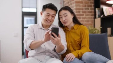 Family time. Multigenerational spouses web surfing on smartphone together, watching social media news, discussing what they see, resting at home