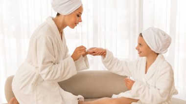 First Manicure Concept. Caring Mother Applying Nail Polish To Her Cute Little Daughter, Mom And Female Child Wearing White Bathrobes Having Beauty Day At Home, Enjoying Self-Care Routine