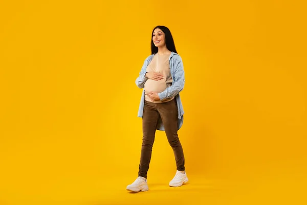 Happy pregnancy concept. Pregnant woman walking and touching belly over yellow studio background, full length shot of positive expectant lady, free space